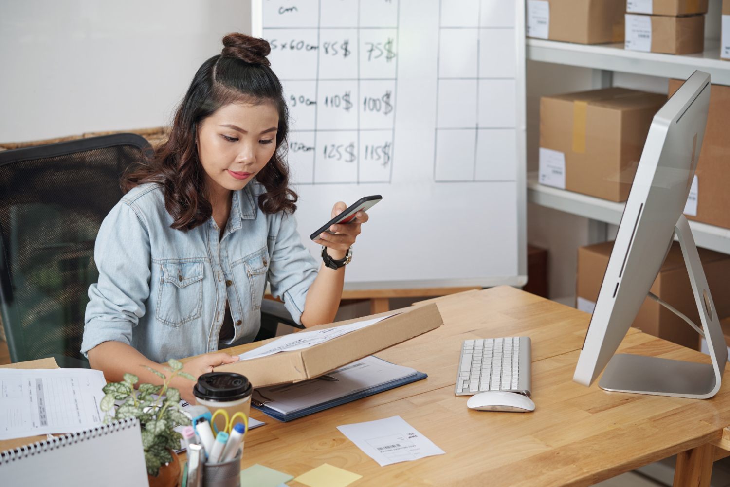 Online seller using a phone while packing a parcel beside a monitor